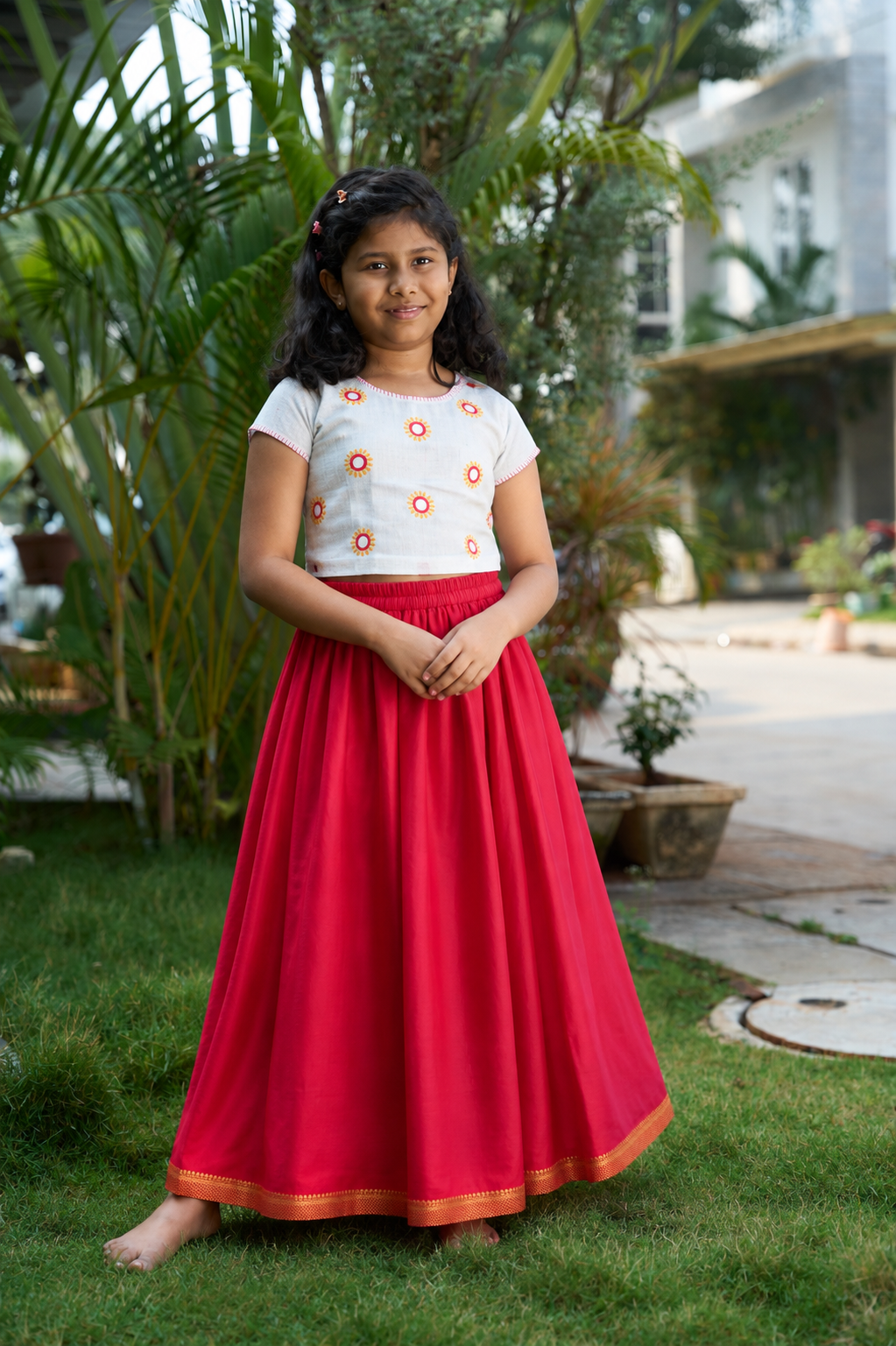 A young girl standing barefoot on a grassy lawn in front of tropical plants and a house, wearing a white short-sleeve top with orange floral embroidery and a long, flowing red skirt with a gold border.
