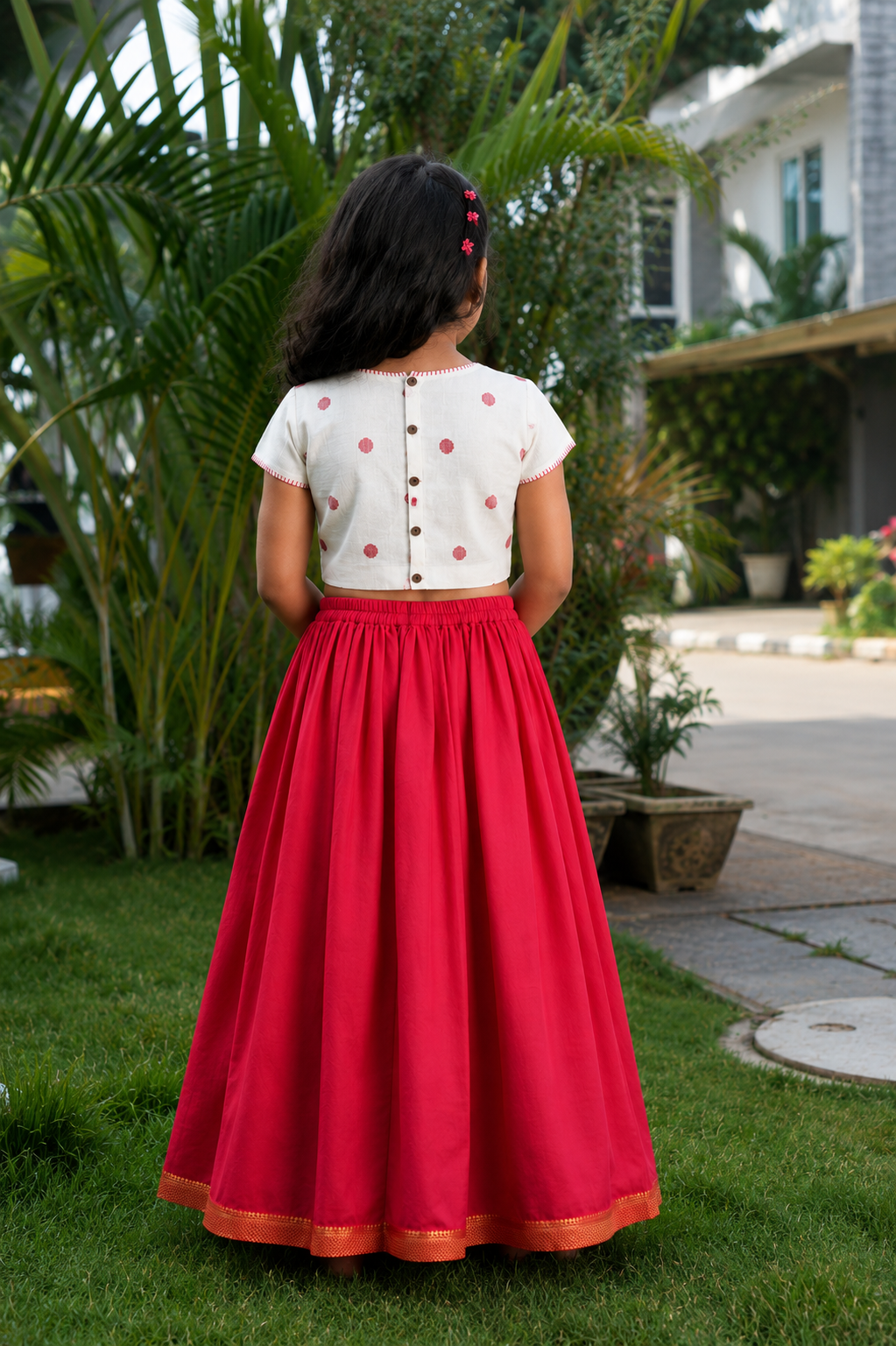 A young girl viewed from behind wearing a traditional Indian outfit consisting of a white short-sleeve blouse with red polka dots and a long red skirt with a gold border, standing on a green lawn in a garden.