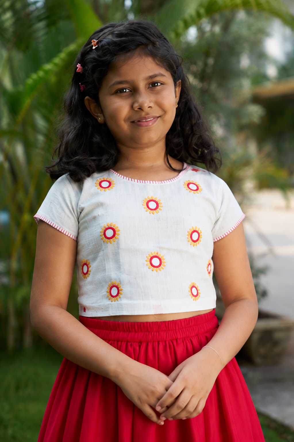 A young girl with dark curly hair wearing a white short-sleeve crop top with red and yellow sunburst patterns, paired with a red skirt, standing outdoors in front of a blurred tropical background.