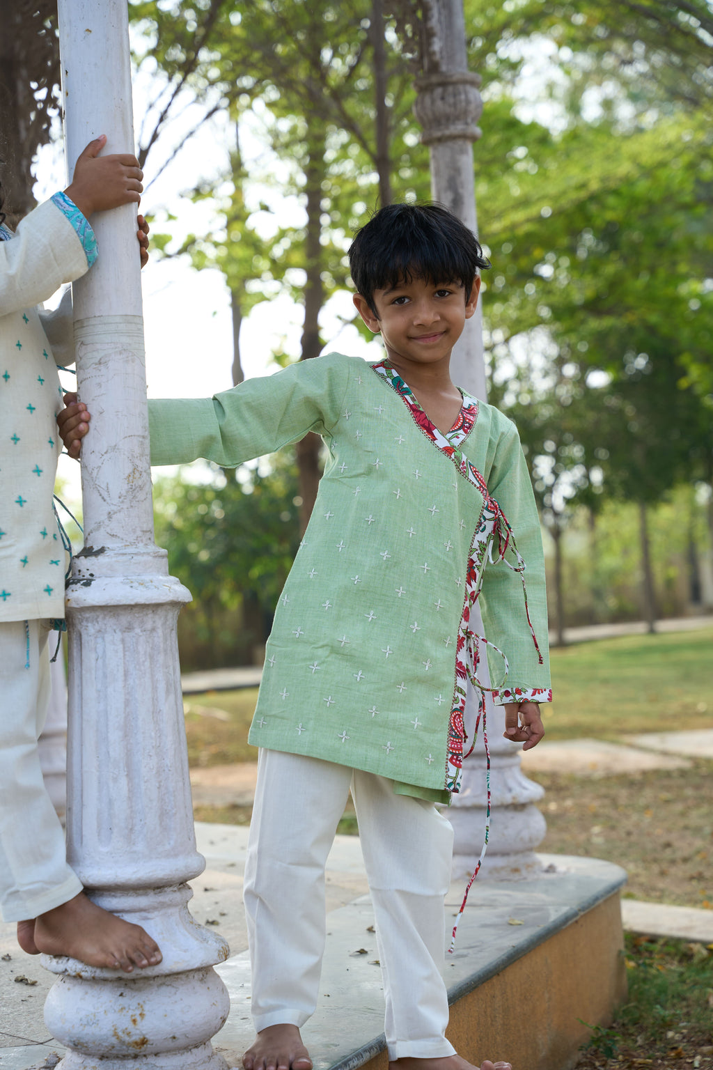 Child in a green kurta with white pants standing outdoors near a decorative column.