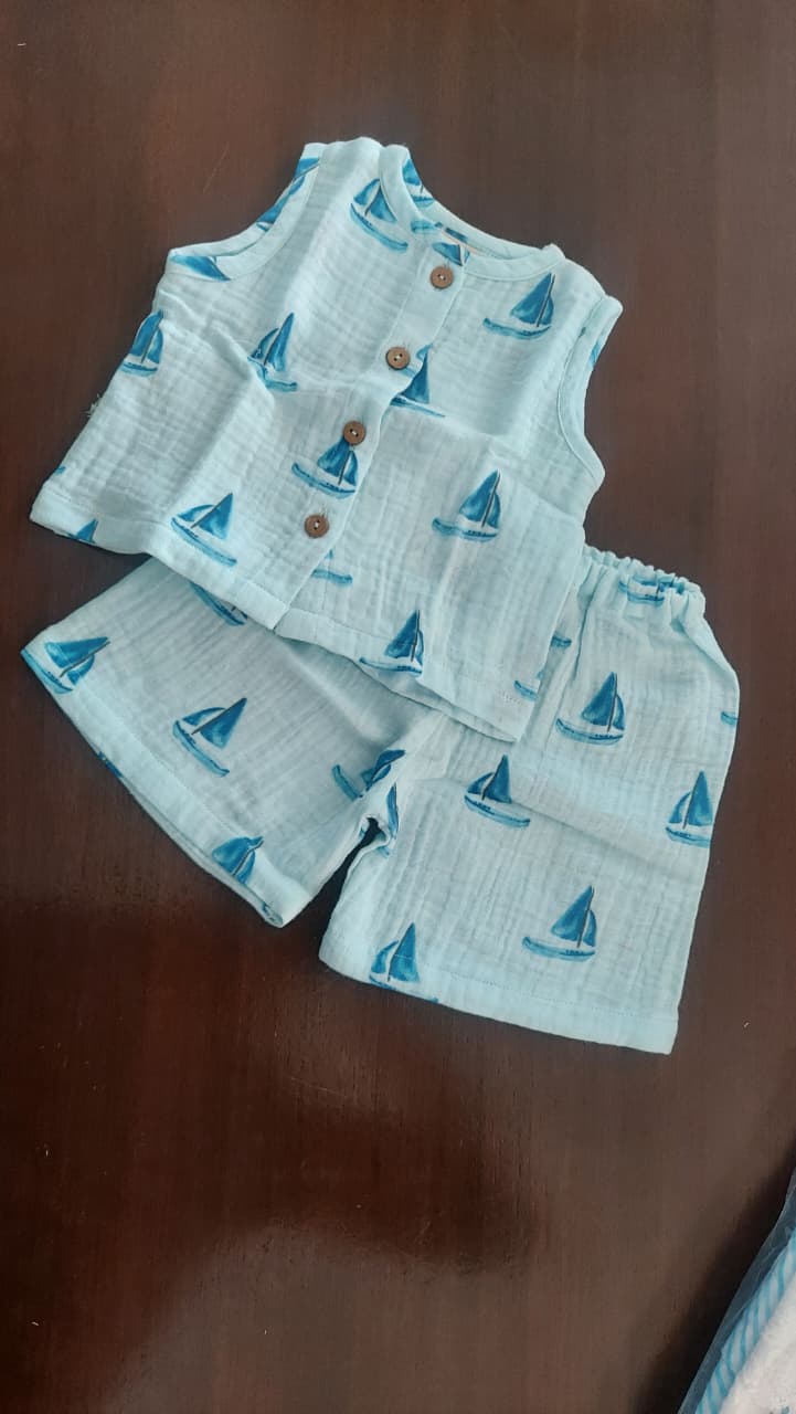 A baby two-piece outfit set consisting of a sleeveless button-front vest and matching shorts, printed with a blue sailboat pattern on a white textured fabric, laid flat on a dark brown wooden surface.