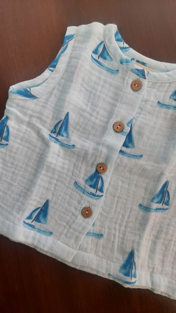 A sleeveless white cotton gauze top with a blue sailboat print and brown wooden buttons, laid flat on a dark brown wooden surface.