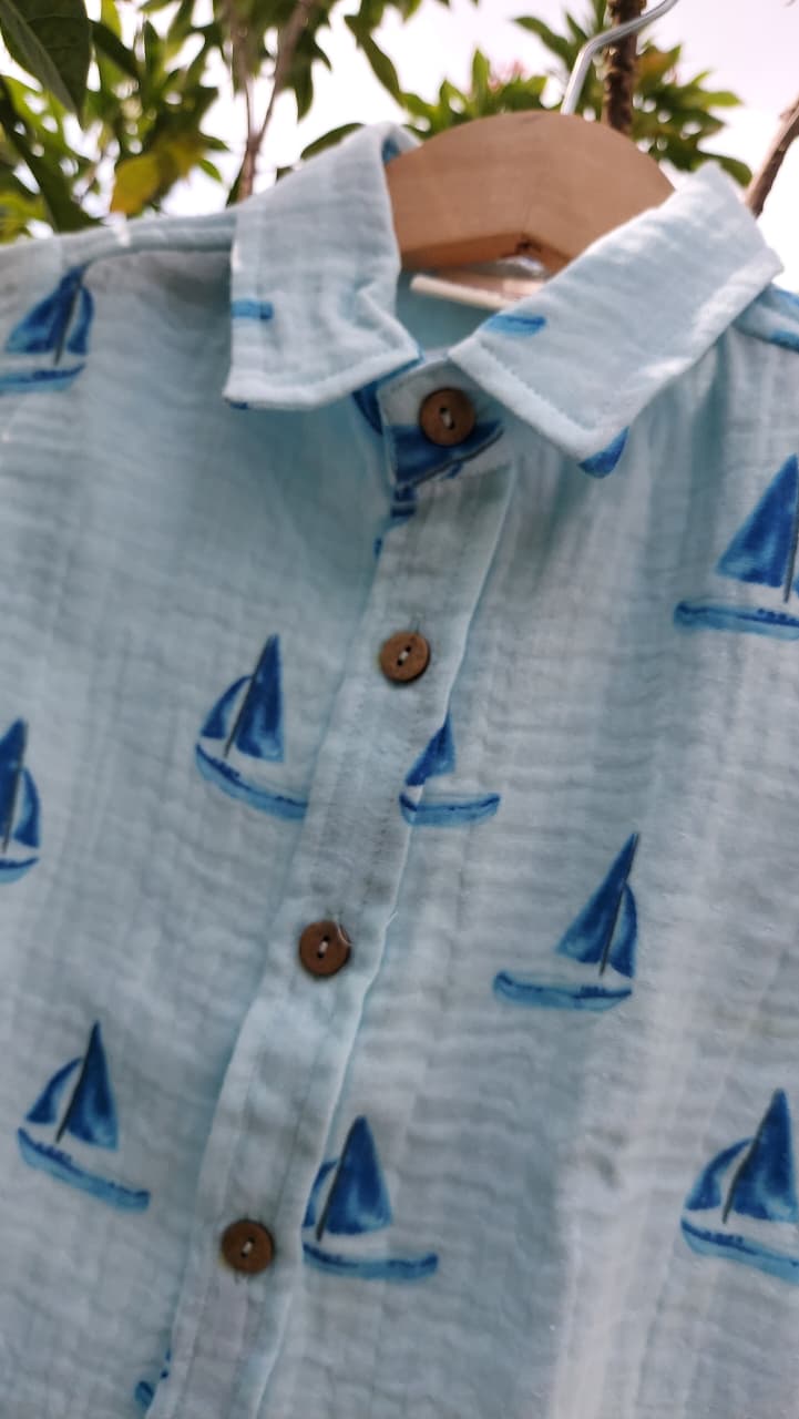 A light blue cotton button-up shirt with a blue sailboat print, hanging on a wooden hanger outdoors.