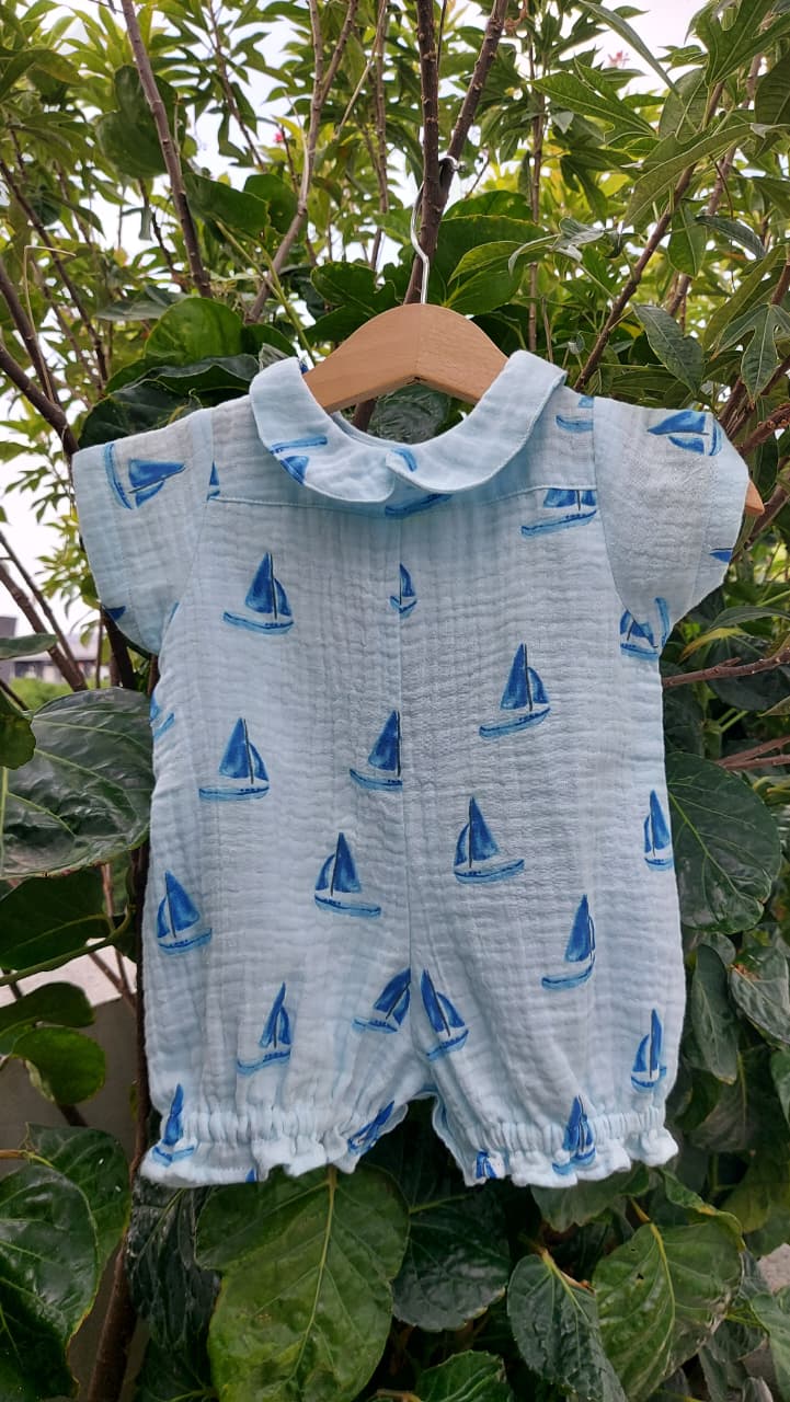 A baby short-sleeve romper in light blue muslin fabric with a blue sailboat print, hanging on a wooden hanger against a background of green garden leaves.