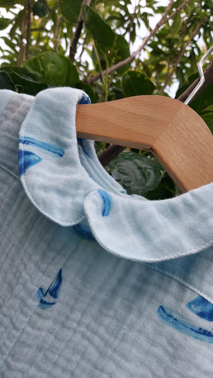 A close-up of a light blue textured fabric garment with a ruffled collar, featuring blue watercolor-style sailboat prints, hanging on a wooden hanger against a background of green leaves.