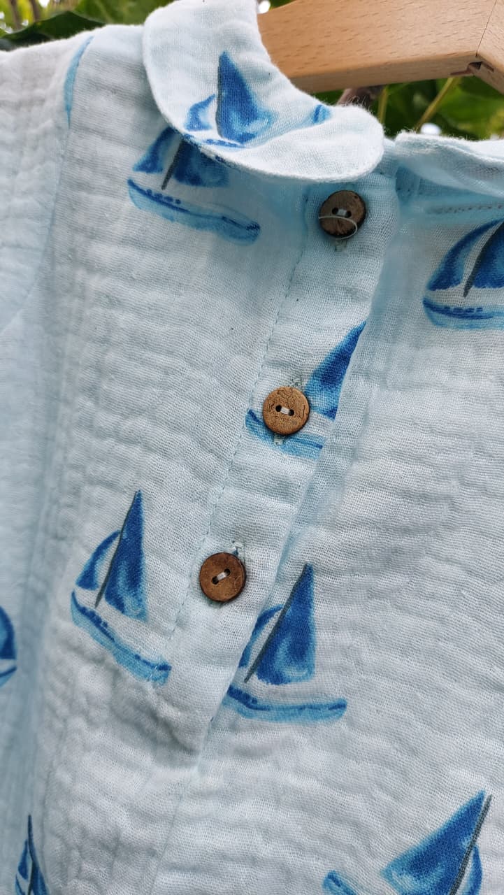 A close-up of a light blue crinkled cotton shirt with a blue sailboat pattern, featuring three brown wooden buttons on the front, hanging on a wooden hanger.