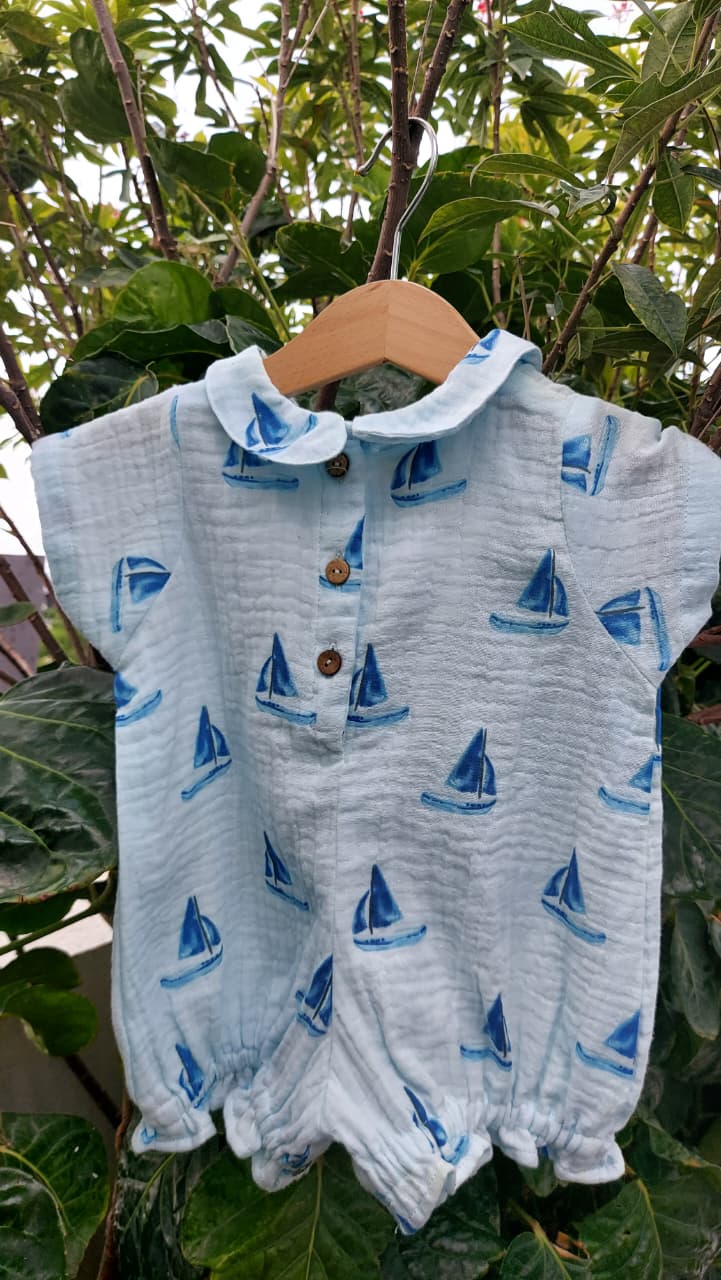 A light blue crinkle gauze baby romper with a blue sailboat print, hanging on a wooden hanger against a background of green garden foliage.
