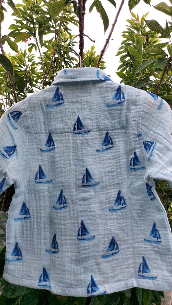 A light blue short-sleeve shirt with a repeating blue sailboat pattern, shown hanging outdoors against a background of green leaves and branches.