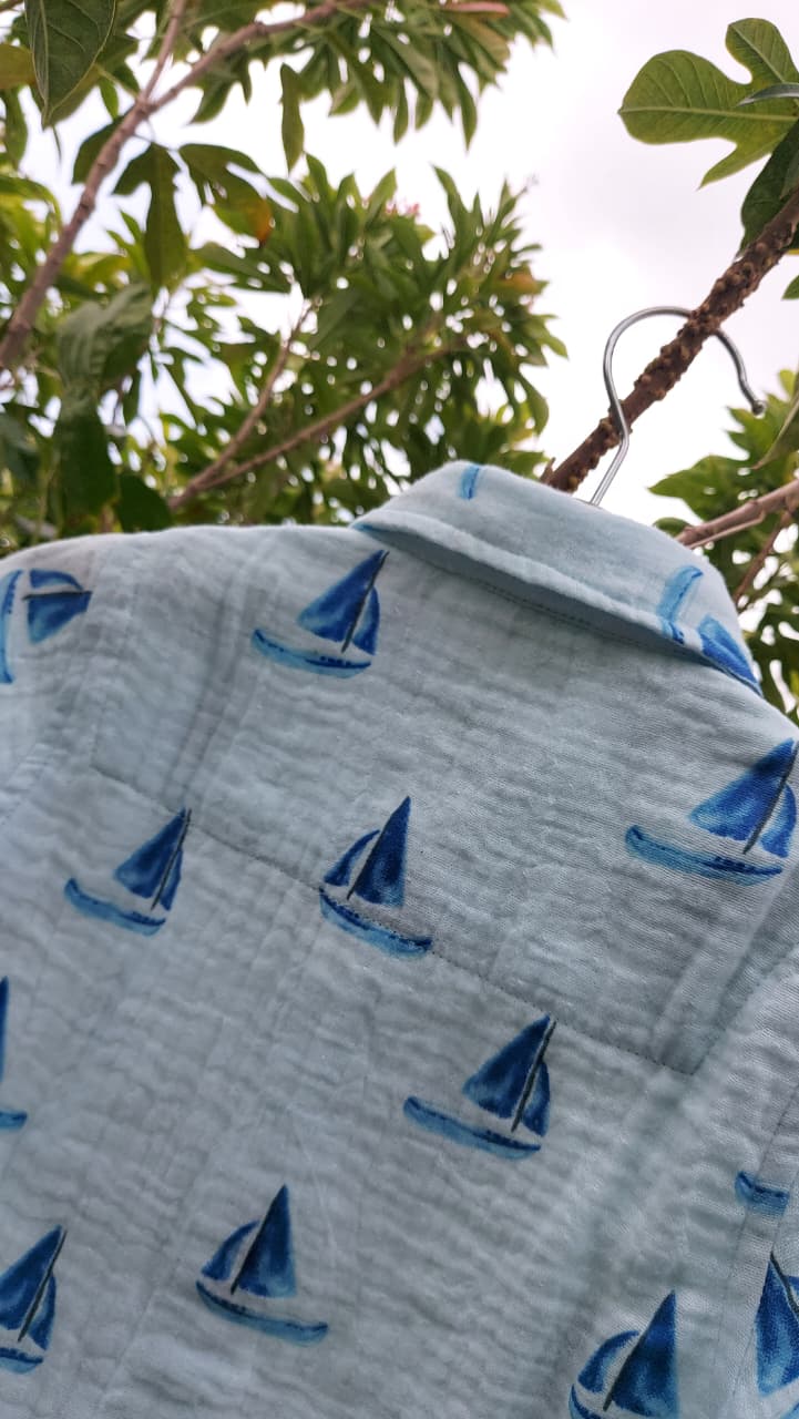 A light blue crinkle-textured fabric item, likely a blanket or swaddle, printed with a repeating pattern of blue and navy sailboats, hanging from a metal hook outdoors against a background of green leaves.