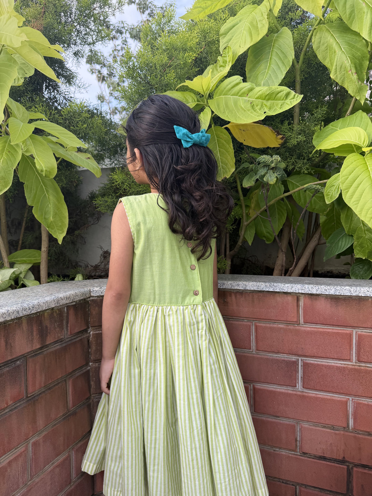 A young girl with long dark curly hair wearing a light green sleeveless dress with a pleated skirt, standing outdoors next to a brick wall with green plants.