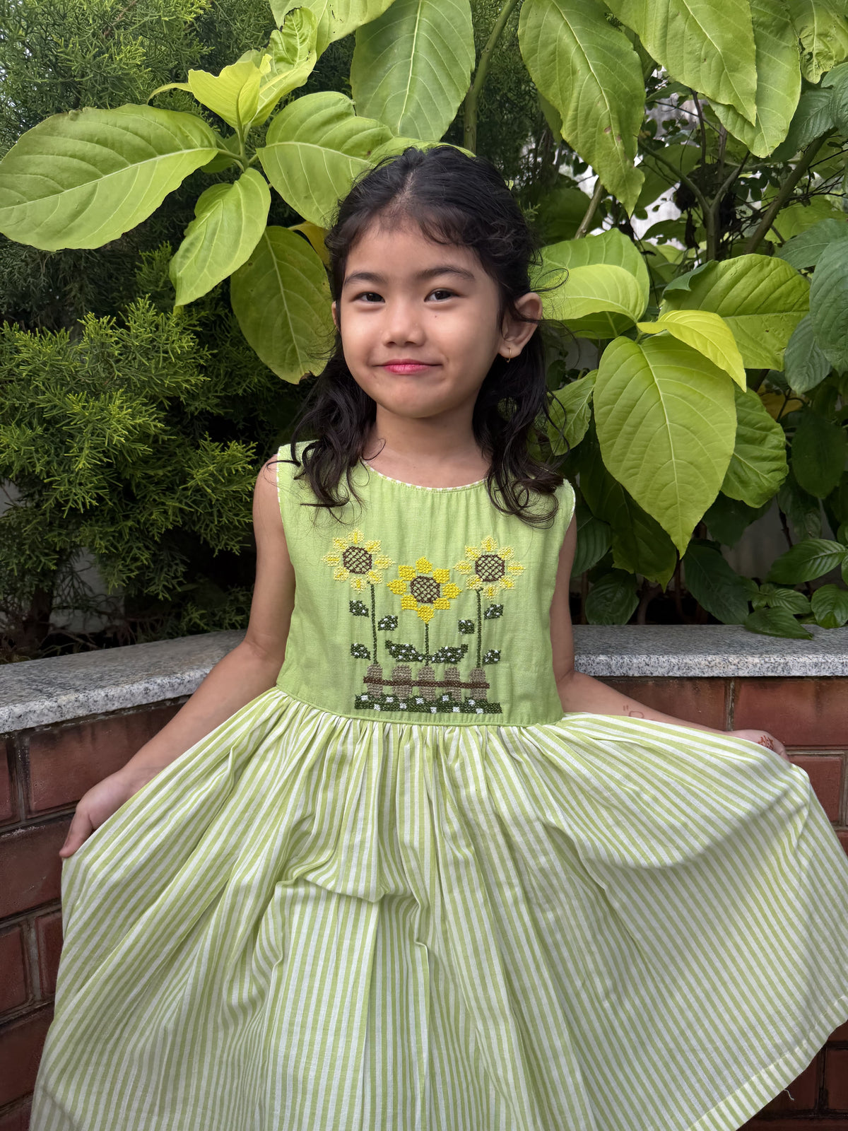 A young girl wearing a sleeveless light green dress with yellow sunflower embroidery on the bodice and green-and-white vertical stripes on the skirt, sitting outdoors in front of green plants.