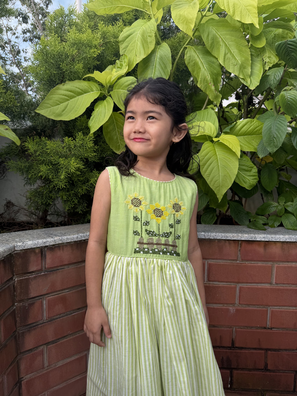 A young girl wearing a light green sleeveless dress with embroidered sunflowers and a smocked bodice, standing outdoors in front of a brick wall and green foliage.