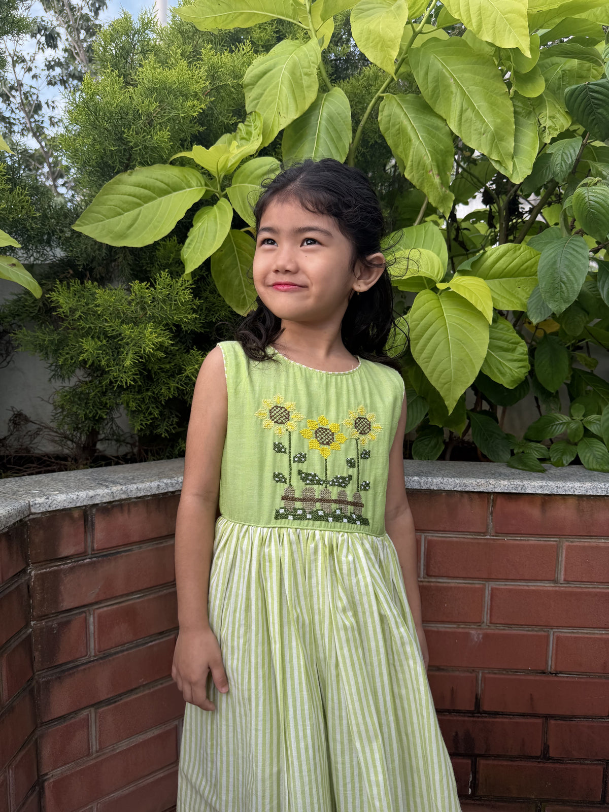 A young girl wearing a light green sleeveless dress with embroidered sunflowers and a smocked bodice, standing outdoors in front of a brick wall and green foliage.