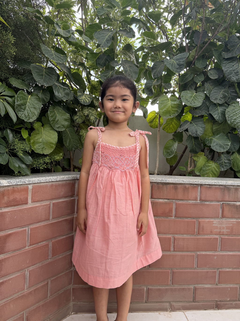 Young girl in a pink dress standing against a brick wall with green foliage.