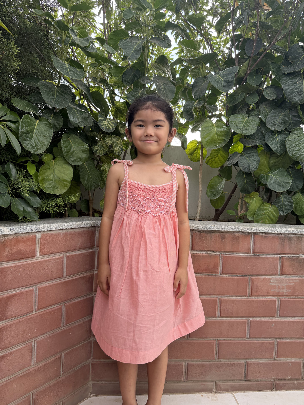 Young girl in a pink dress standing against a brick wall with green foliage.