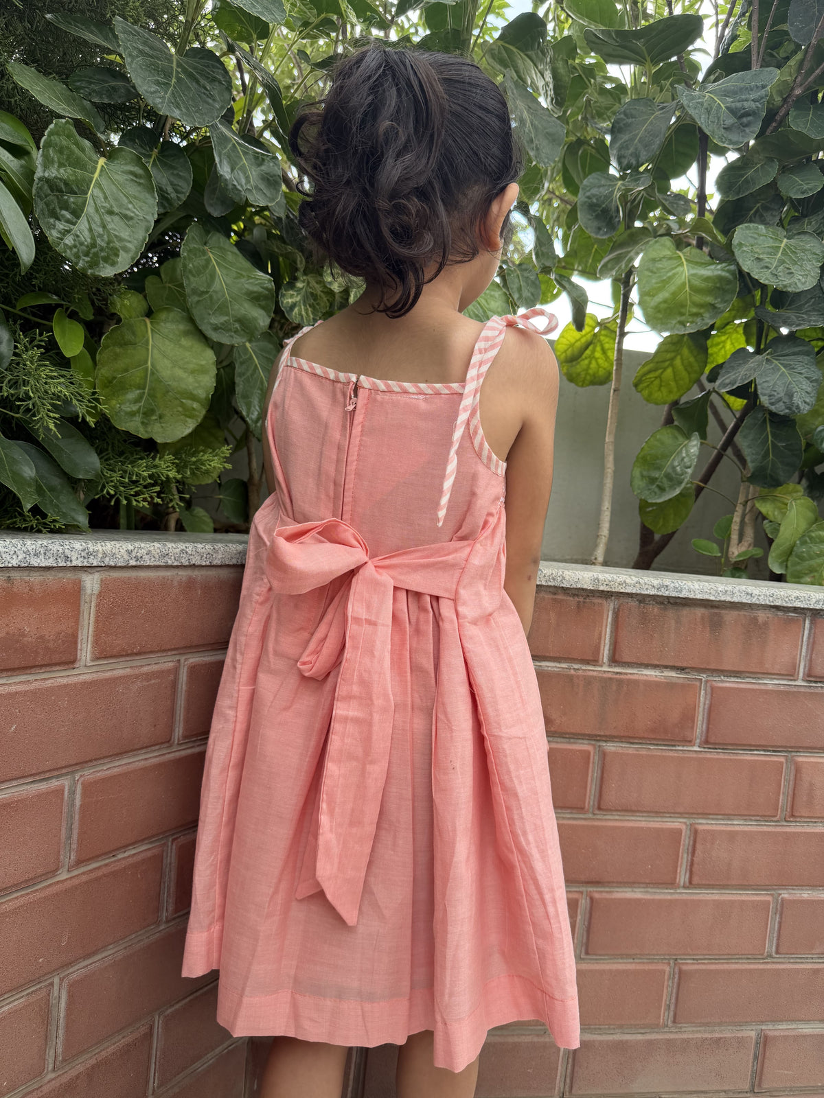 Person wearing a pink dress with a bow tie standing against a brick wall with green foliage.