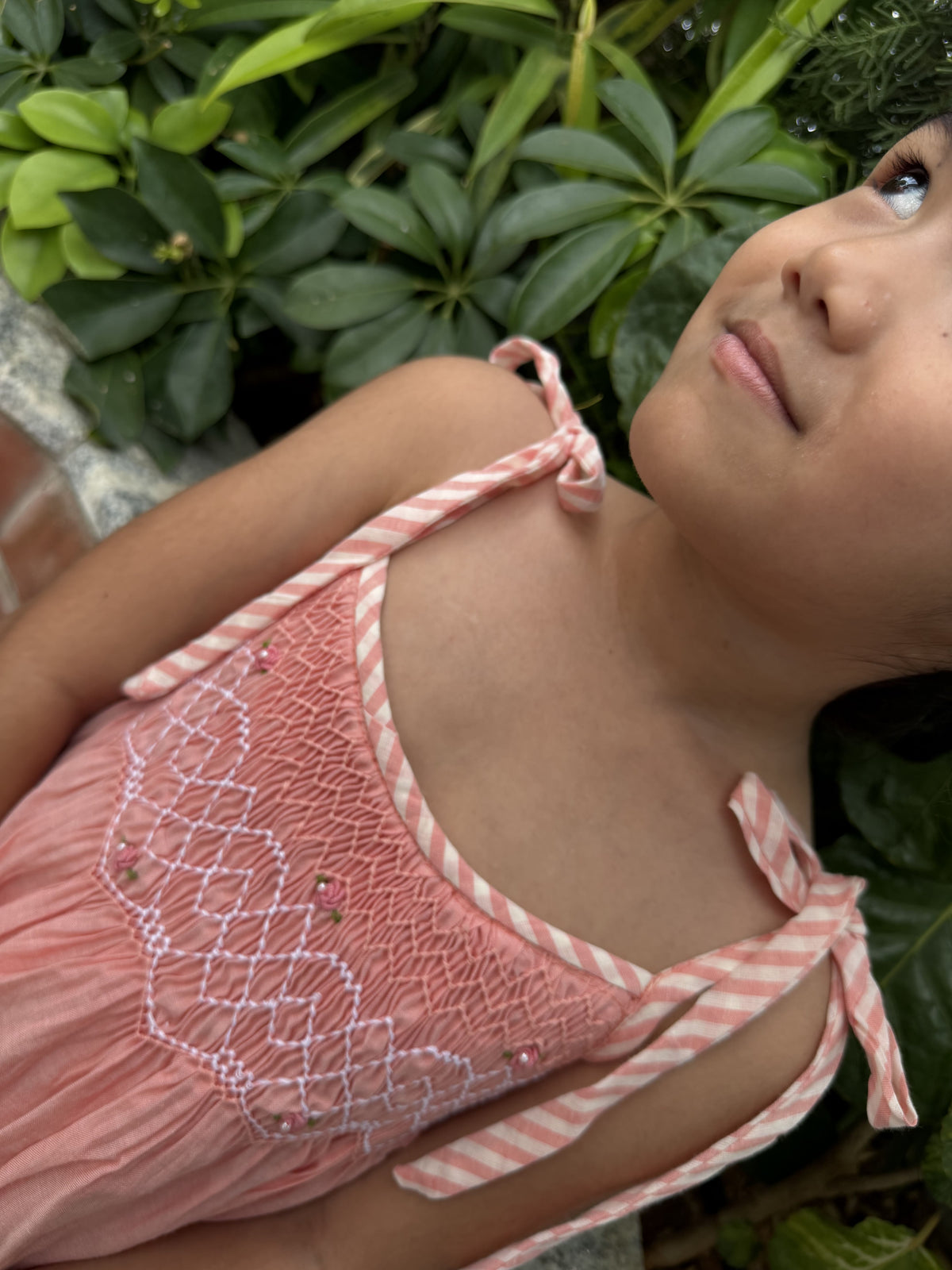 Child wearing a pink and white striped dress with floral details, standing among green foliage.