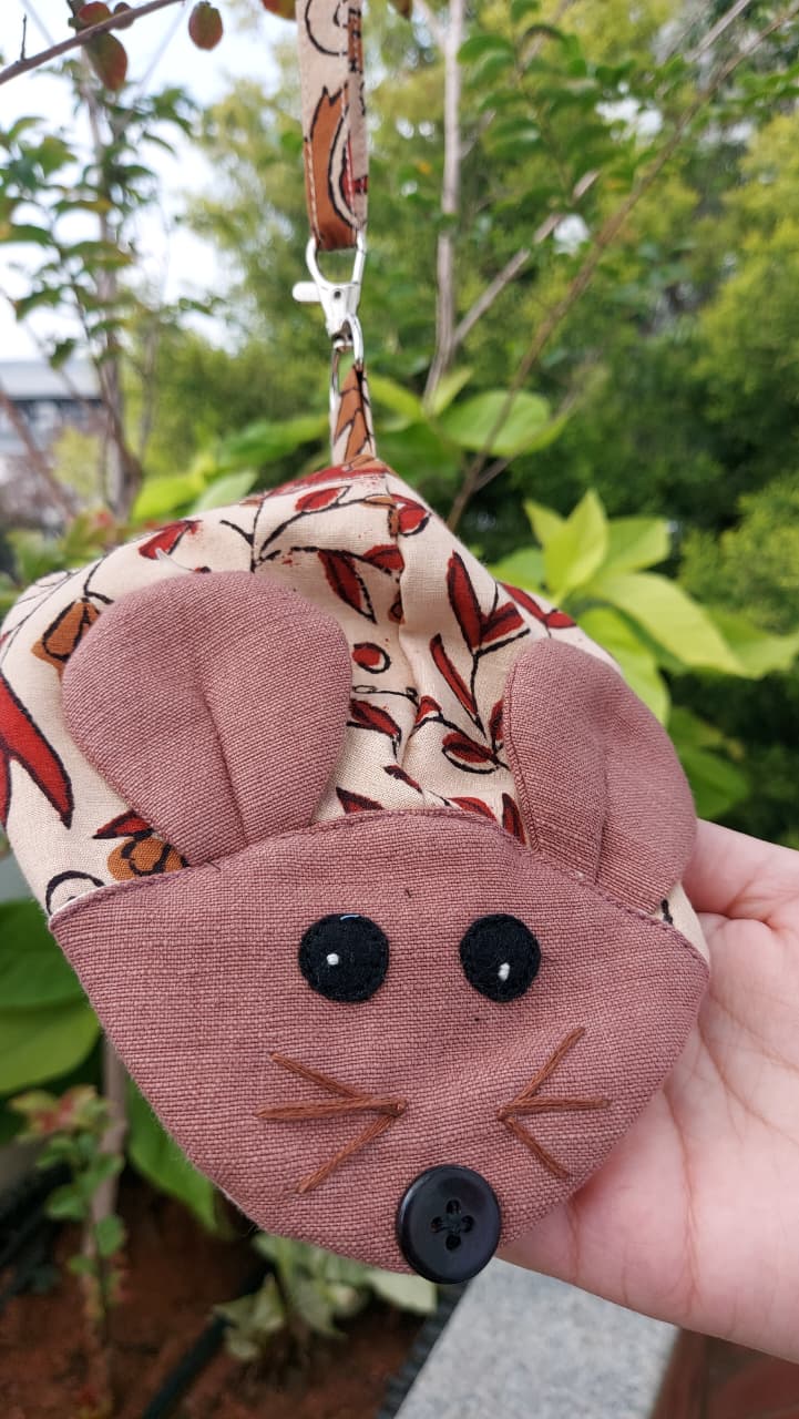 A handmade mouse-shaped fabric pouch held in a hand outdoors. The pouch features a dusty pink fabric face with embroidered whiskers and a black button nose, a patterned fabric top with red and brown motifs, and a matching patterned wrist strap.