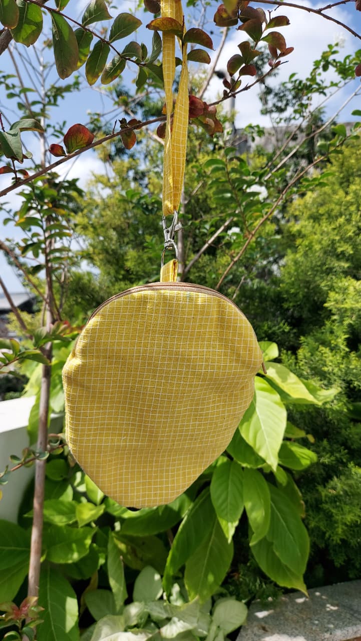 A small yellow fabric pouch with a grid pattern and a matching hanging ribbon, photographed outdoors among green leaves and branches.