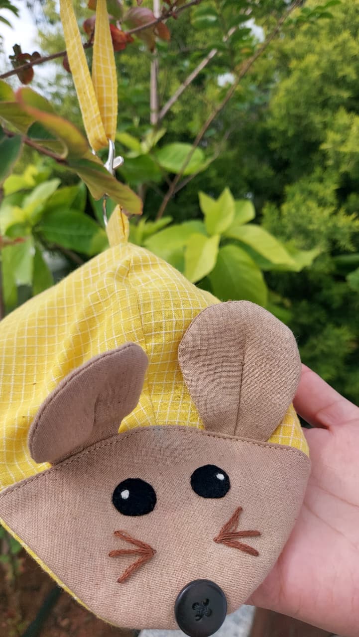A small yellow and white checkered fabric mouse-shaped pouch or bag being held by a hand outdoors, with green foliage in the background.