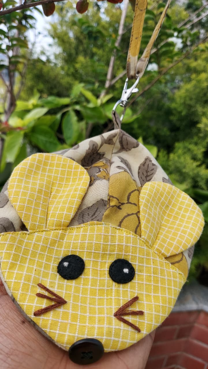 A handmade yellow mouse-shaped fabric pouch with embroidered facial features and a metal clasp, held in a hand against a blurred green garden background.
