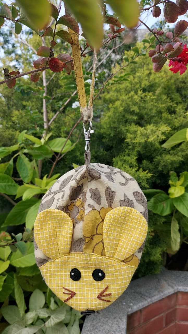 A handmade fabric pouch shaped like a mouse head, featuring a yellow grid-patterned face and ears, black bead eyes, and a brown stitched nose and whiskers, hanging from a measuring tape strap against a background of green garden foliage.