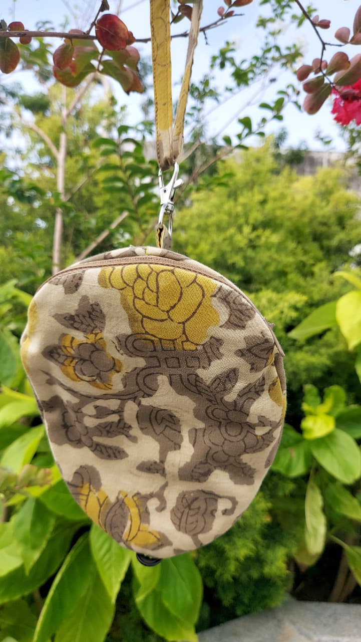 A small fabric pouch with a floral and dragon pattern in yellow, brown, and beige, hanging from a strap outdoors in front of green foliage.