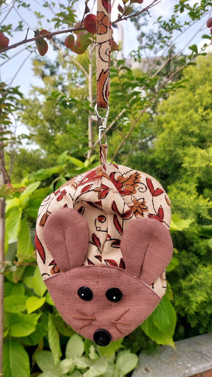 A small fabric mouse-shaped pouch or bag hanging from a leafy branch outdoors. The pouch has a floral print in beige, brown, and muted pink, with a solid pink mouse face, black button eyes, and a matching floral wrist strap.