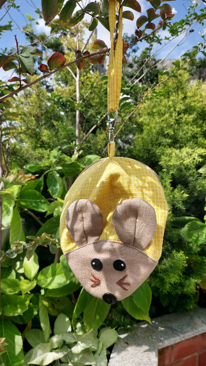 A yellow mesh mouse-shaped hanging bag with a beige fabric face, black embroidered eyes and nose, and a yellow ribbon loop, photographed outdoors among green leaves and branches.