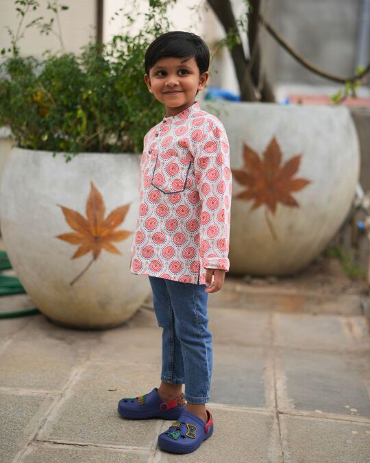 Child wearing a patterned shirt and blue jeans standing outdoors with decorative pots in the background.